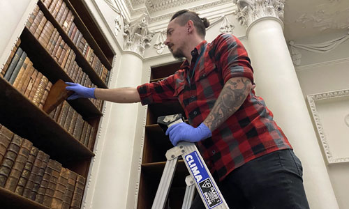 A male volunteer is stood on some step ladders looking through books in the Georgian Library of Temple Newsam House