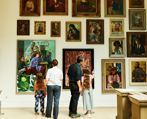 A family of four look at a variety of brightly coloured portrait paintings hung on a wall in a gallery. They include works by artists including Pablo Picasso and Paula Rego.