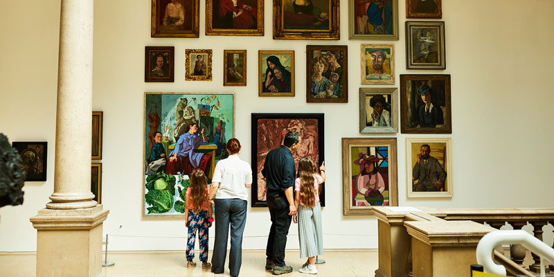 A family of four look at a variety of brightly coloured portrait paintings hung on a wall in a gallery. They include works by artists including Pablo Picasso and Paula Rego.