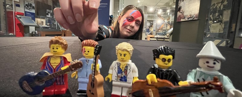 Lego figurines of David Bowie. A person in the background is wearing Bowie style makeup and reaching for the figurines.