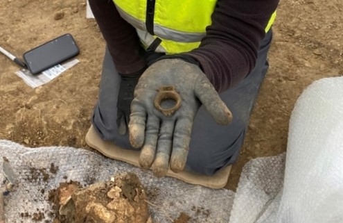 A person kneels on brown earth holding a ring in their gloved hand