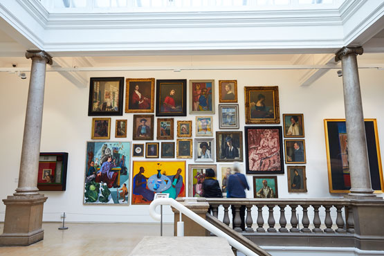 A collection of portrait paintings is hung in a gallery by a Victorian staircase. It is being observed by two women