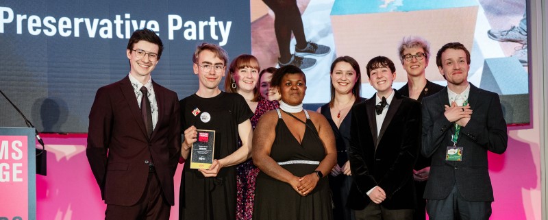 A group of young people standing on a stage smiling and holding an award trophy. A screen behind them reads: Preservative Party
