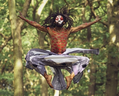 a man with red and white paint on his face and torso jumps in the air with trees in the background