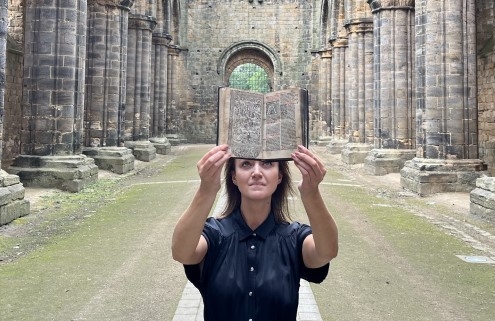 A woman holds up an old book in the ruins of Kirkstall Abbey