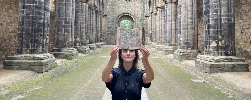 A woman holds up an old book in the ruins of Kirkstall Abbey