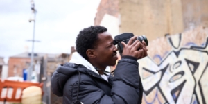 A young male holding a filming camera to his eye, looking at a walk with graffiti on