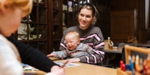 a woman smiling and holding a baby with craft materials on the table in front of them