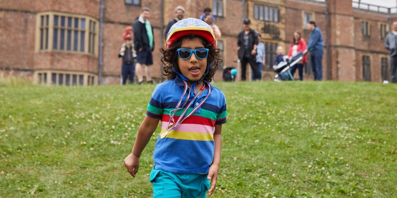 running child wearing sunglasses, cycling helmet and gold medal with temple newsam house in the background