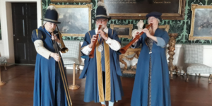 three people dressed in historical costumes and hats, one playing a trombone and two playing wind instruments