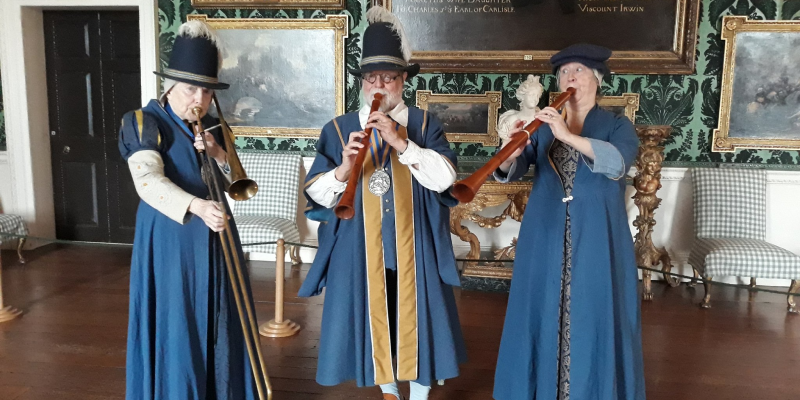 three people dressed in historical costumes and hats, one playing a trombone and two playing wind instruments